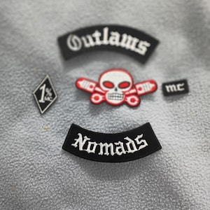 以下が含まれることがあります： 黒と白の刺繍パッチのコレクション。パッチには、カーブしたデザインの「Outlaws」という単語、交差したピストンが付いたスカル、「Nomads」という単語、数字の「1%」、文字の「mc」が含まれています。