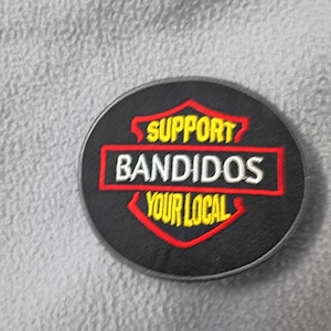May include: A round black patch with the words "SUPPORT BANDIDOS YOUR LOCAL" in white and red lettering. The text is inside a red and yellow shield design. The patch has a black border and is on a gray, textured surface.