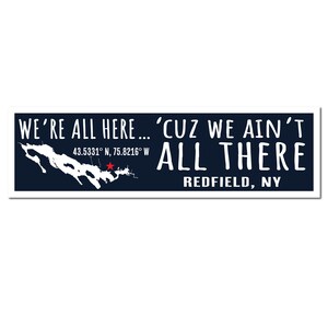 Op de afbeelding: Een marineblauw rechthoekig bord met witte tekst met de tekst "WE'RE ALL HERE... 'CUZ WE AIN'T ALL THERE REDFIELD, NY." Links staat een witte kaart van een meer met een rode ster. Coördinaten worden vermeld.
