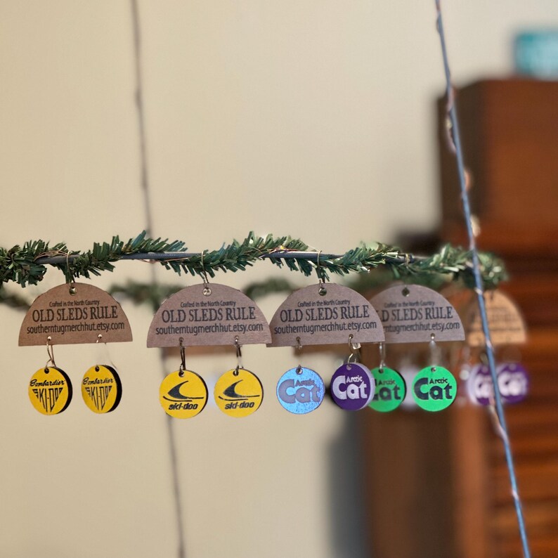 Puede incluir: Una exhibici&oacute;n de pendientes de madera con varios dise&ntilde;os y texto. Los pendientes est&aacute;n suspendidos de una guirnalda de vegetaci&oacute;n. Los dise&ntilde;os incluyen el texto "OLD SLEDS RULE" y logotipos de "Bombardier Ski-Doo" y "Arctic Cat".