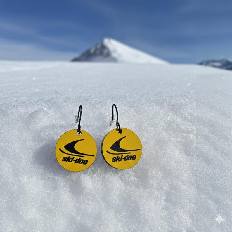 Puede incluir: Un par de pendientes redondos amarillos con un logotipo negro de ski-doo, colgando sobre un fondo de monta&ntilde;a nevada. Los pendientes tienen ganchos negros y el logotipo de ski-doo es negro.