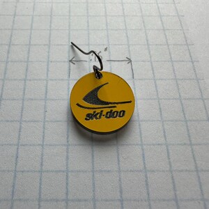 Puede incluir: Un pendiente circular amarillo con el logotipo negro de ski-doo. El pendiente tiene un gancho negro y mide aproximadamente 1,9 cm de di&aacute;metro. El logotipo de ski-doo presenta una ola negra estilizada.