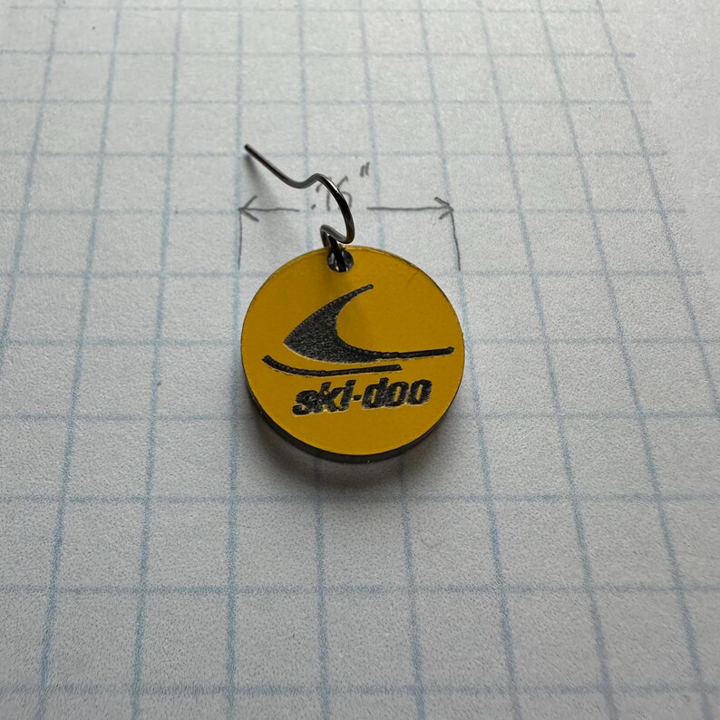 Puede incluir: Un pendiente circular amarillo con el logotipo negro de ski-doo. El pendiente tiene un gancho negro y mide aproximadamente 1,9 cm de di&aacute;metro. El logotipo de ski-doo presenta una ola negra estilizada.