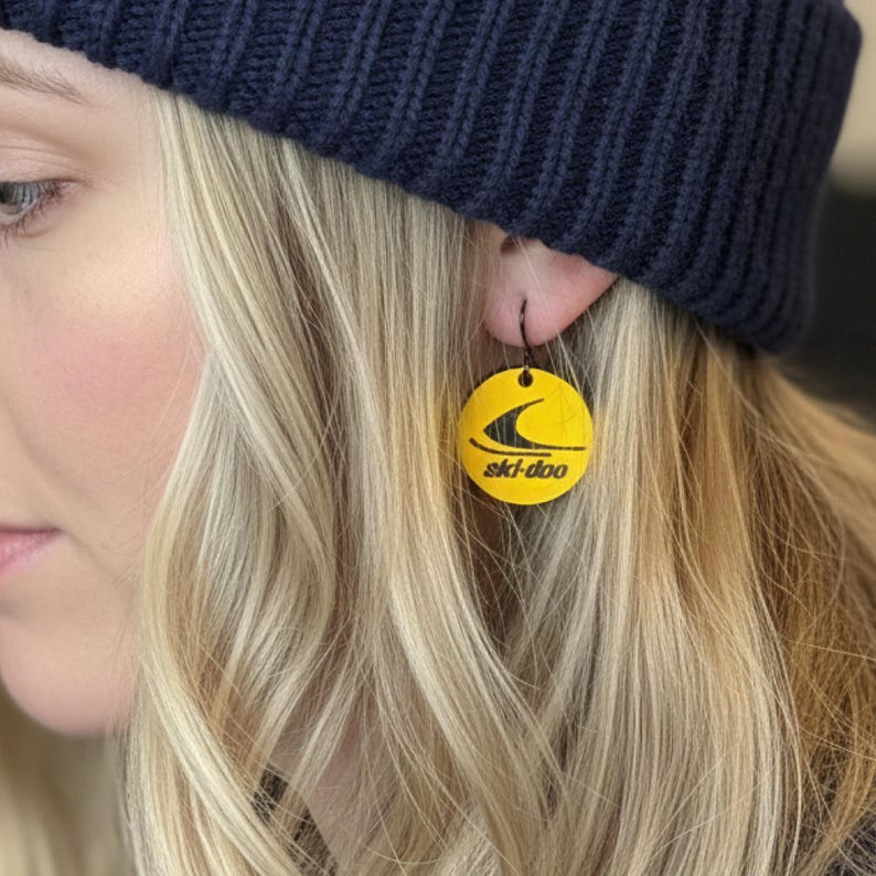 Puede incluir: Pendientes redondos amarillos con el logotipo negro de Ski-Doo. Los pendientes cuelgan de ganchos negros. Los pendientes los lleva una persona con pelo rubio y un gorro azul marino.