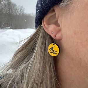 Puede incluir: Pendientes redondos amarillos y negros con el texto "ski-doo" y un gr&aacute;fico estilizado. Los pendientes cuelgan de un gancho negro. El fondo es una escena nevada al aire libre.
