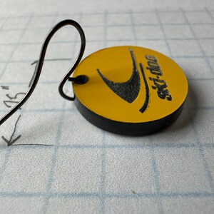 Puede incluir: Un pendiente de metal negro con un dije circular amarillo. El dije tiene un logotipo negro y el texto "ski-doo". La longitud total del pendiente es de aproximadamente 4,4 cm.