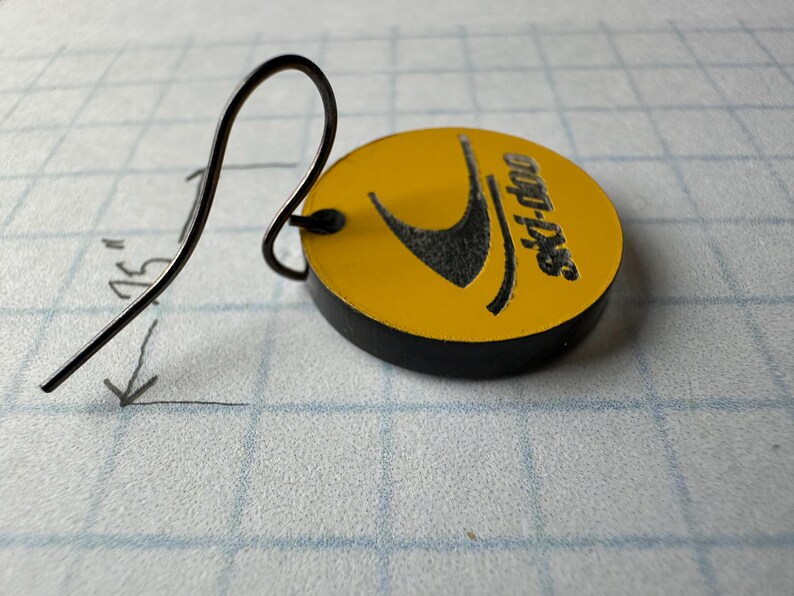 Puede incluir: Un pendiente de metal negro con un dije circular amarillo. El dije tiene un logotipo negro y el texto "ski-doo". La longitud total del pendiente es de aproximadamente 4,4 cm.