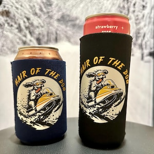 May include: Two beverage insulators, one navy blue and one black, each with a circular graphic. The graphic features a dog wearing goggles on a snowmobile, with the text "HAIR OF THE DOG" in gold. One insulator holds a can.