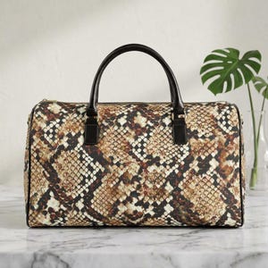 May include: A rectangular duffel bag with a snakeskin pattern in shades of brown, tan, and black. The bag has black leather handles and accents. The bag is sitting on a marble surface.