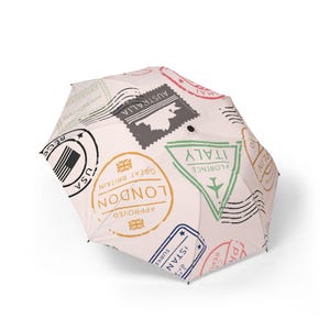 May include: A white umbrella with a repeating pattern of travel-themed stamps. The stamps include the USA, London, Australia, and Italy. The umbrella is open and angled, showcasing the design.