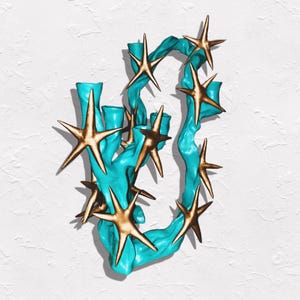 May include: A teal-colored, abstract sculpture with gold-colored starfish accents. The sculpture has a textured, organic shape, with the starfish seemingly attached to the teal form. The background is a textured white surface.