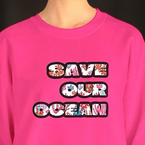May include: A vibrant pink sweatshirt featuring the text "SAVE OUR OCEAN". The letters are white with a black border and filled with a colourful coral reef pattern.