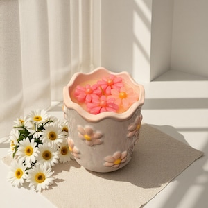 May include: A ceramic flower pot with a wavy rim, decorated with embossed daisy details. The pot contains pink flower-shaped candles floating in a liquid. A bouquet of daisies sits beside the pot on a neutral-coloured cloth, creating a floral theme.