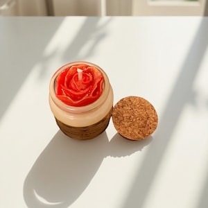 Puede incluir: Una vela roja con forma de rosa en un tarro de cristal transparente con tapa de corcho. La vela tiene una mecha blanca y está en un tarro con una banda marrón. La tapa de corcho está al lado del tarro sobre una superficie blanca.