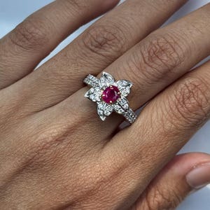 Könnte beinhalten: Ein silberfarbener Ring mit einem zentralen, oval geschliffenen roten Rubin, der von einer Ansammlung kleiner, klarer, rund geschliffener weißer Diamanten in Blütenblattform umgeben ist, mit weiteren kleinen Diamanten entlang der Ringschiene.