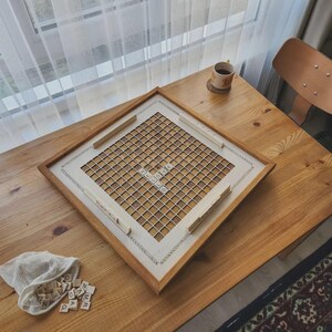 May include: A wooden framed Scrabble board displaying the words "HELLO LOVE". The board sits on a wooden table, accompanied by a small bag of letter tiles and a cup of coffee. A decorative item for the home.
