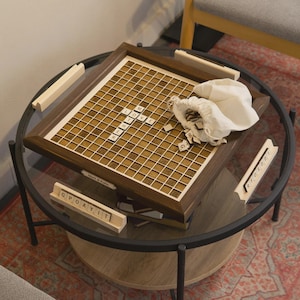 May include: A wooden Scrabble board with the word "LOVE" spelled out in tiles. The board is brown with a grid pattern and sits on a glass-topped table. A small white bag and additional tiles are nearby. The words "Mark & Kate" are engraved on the board.