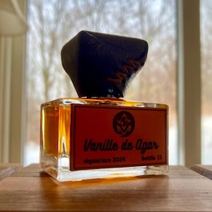 May include: A square glass perfume bottle with a dark brown cap. The label reads "Vanille de Agar" with "signature 2024" and "bottle 11" below. The liquid inside is amber-colored. The bottle sits on a wooden surface.