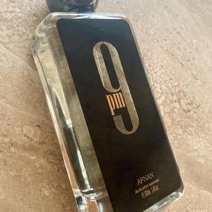 May include: Clear rectangular perfume bottle with a black label featuring the text "9 pm" in a stylized font. The bottle has a dark, spherical cap and contains a clear liquid. The brand name "AFNAN" is also visible.