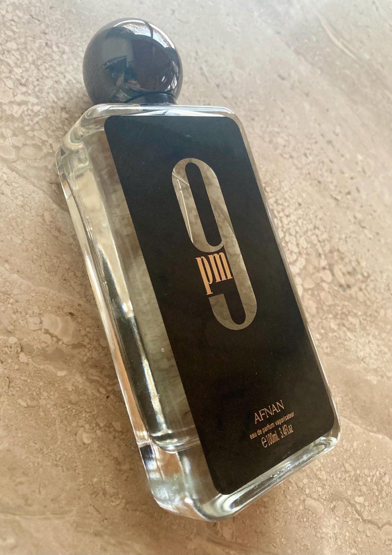 May include: Clear rectangular perfume bottle with a black label featuring the text "9 pm" in a stylized font. The bottle has a dark, spherical cap and contains a clear liquid. The brand name "AFNAN" is also visible.