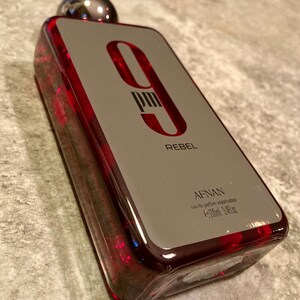 May include: A rectangular perfume bottle with a silver cap and a red-tinted glass body. The label is gray with the text "9 PM REBEL" in red. The bottle contains 3.4 fl oz of perfume.