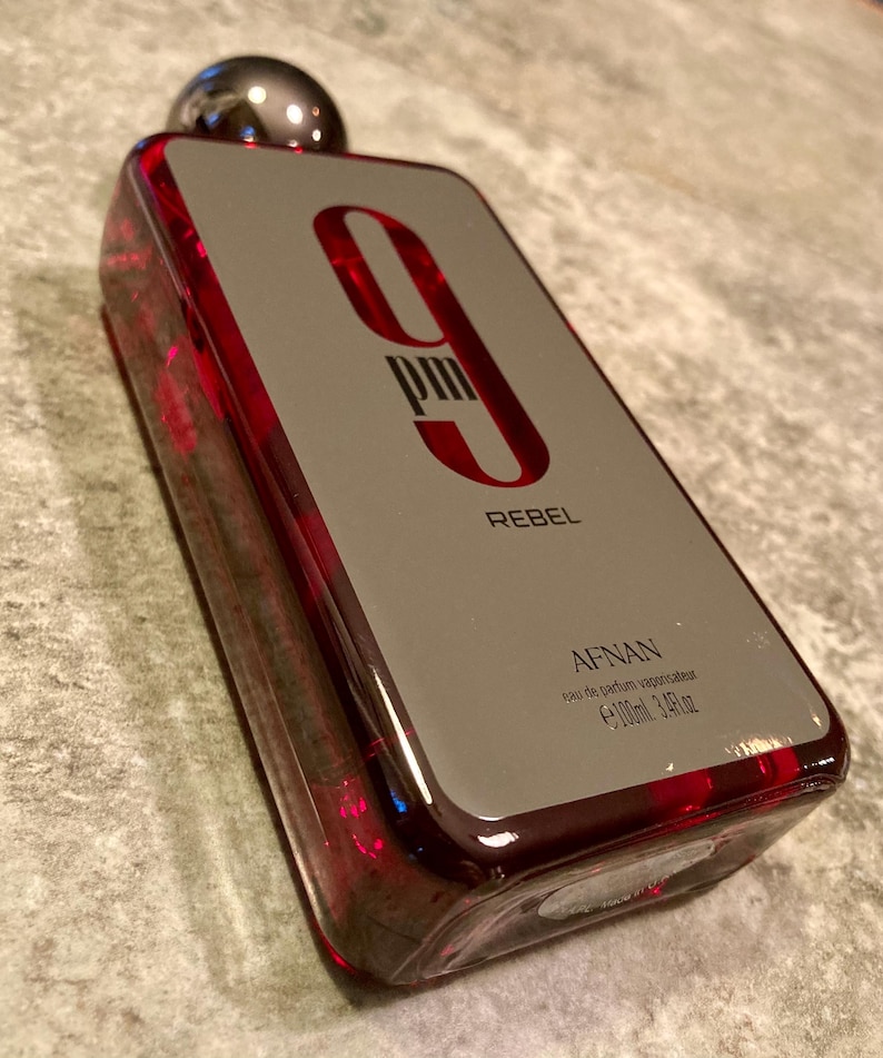 May include: A rectangular perfume bottle with a silver cap and a red-tinted glass body. The label is gray with the text "9 PM REBEL" in red. The bottle contains 3.4 fl oz of perfume.