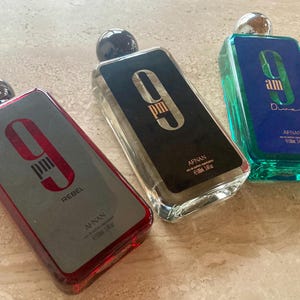 May include: Three rectangular perfume bottles with silver caps. One bottle is red with "9 PM Rebel" text, another is black with "9 PM" text, and the third is blue-green with "am Dive" text. Each bottle has a silver cap and is labeled "AFNAN".