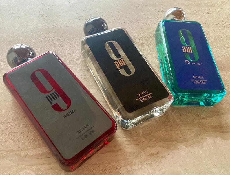 May include: Three rectangular perfume bottles with silver caps. One bottle is red with "9 PM Rebel" text, another is black with "9 PM" text, and the third is blue-green with "am Dive" text. Each bottle has a silver cap and is labeled "AFNAN".