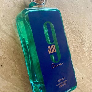 May include: A rectangular perfume bottle with a silver cap. The bottle is filled with a turquoise liquid and has a blue label with the text "9 am Dive" in gold lettering. The brand name "Afnan" is also visible.