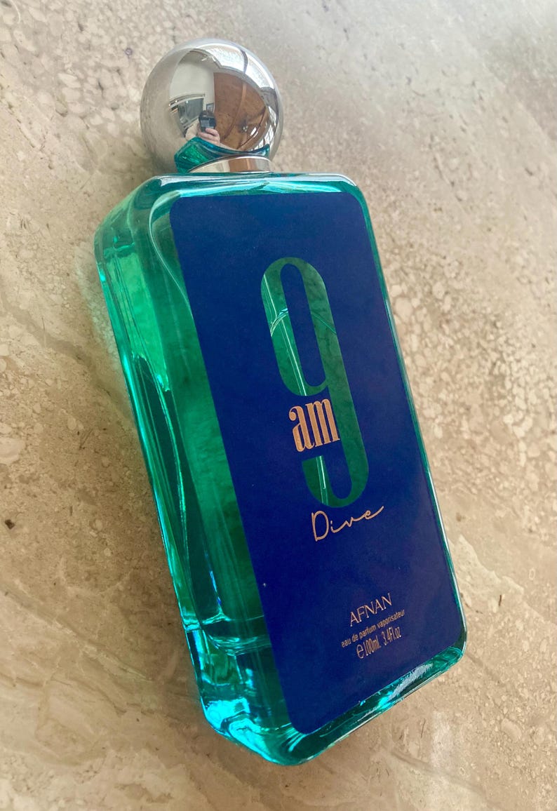 May include: A rectangular perfume bottle with a silver cap. The bottle is filled with a turquoise liquid and has a blue label with the text "9 am Dive" in gold lettering. The brand name "Afnan" is also visible.
