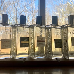 May include: Five clear glass cologne bottles with black caps are arranged on a wooden surface. Each bottle has a rectangular shape and a label with the brand name "CREMO" and a fragrance name. The bottles are filled with a light amber liquid.