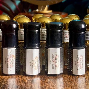 May include: A collection of small perfume bottles with black caps and gold tops. Each bottle has a label with text, including "Sicilian Vanilla Attar 2021" and "Dark Forest 2012 Attar". The bottles are arranged on a wooden surface.
