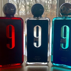 May include: Three rectangular perfume bottles with black spherical caps. The bottles are red, black, and teal, each with a large number "9" on the front. The black bottle has "9 pm" printed on it, and the teal bottle has "9 am Dive" printed on it.