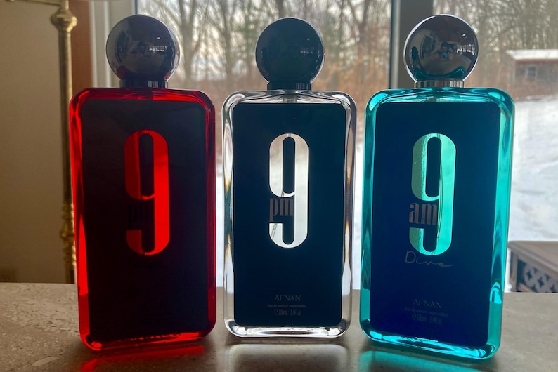 May include: Three rectangular perfume bottles with black spherical caps. The bottles are red, black, and teal, each with a large number "9" on the front. The black bottle has "9 pm" printed on it, and the teal bottle has "9 am Dive" printed on it.