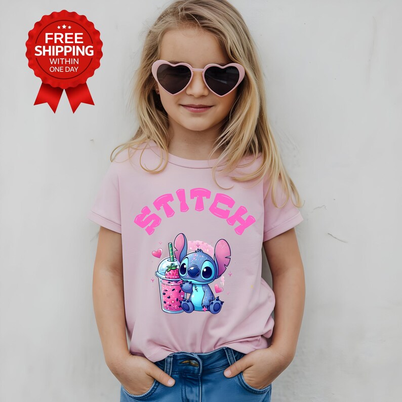 K&ouml;nnte beinhalten: Hellrosa T-Shirt mit dem Wort "STITCH" in rosa Buchstaben. Das Shirt zeigt eine Cartoon-Abbildung von Stitch mit einem rosa Getr&auml;nk. Die Person auf dem Bild tr&auml;gt herzf&ouml;rmige Sonnenbrillen.