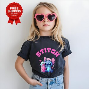 K&ouml;nnte beinhalten: Ein Kind tr&auml;gt ein schwarzes T-Shirt mit dem Wort "Stitch" in Rosa. Das Shirt zeigt eine Zeichentrickfigur, die ein rosa Getr&auml;nk h&auml;lt. Das Kind tr&auml;gt auch rosa herzf&ouml;rmige Sonnenbrillen und blaue Jeans.