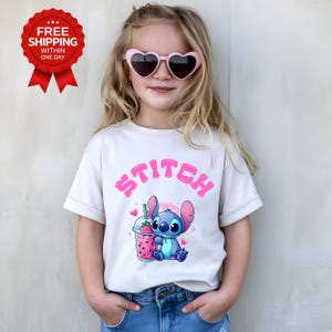 K&ouml;nnte beinhalten: Hellbeige T-Shirt mit dem Wort "STITCH" in rosa Buchstaben &uuml;ber einer Cartoonfigur, die ein rosa Getr&auml;nk mit Herzen h&auml;lt. Die Figur ist blau mit gro&szlig;en Ohren. Die Person tr&auml;gt rosa herzf&ouml;rmige Sonnenbrillen.