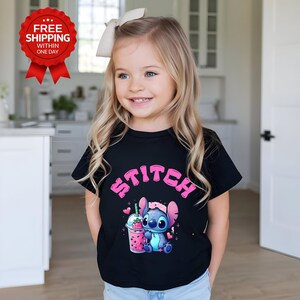 K&ouml;nnte beinhalten: Ein schwarzes T-Shirt mit dem Wort "STITCH" in rosa Buchstaben. Das T-Shirt zeigt eine Zeichentrickfigur, die ein Getr&auml;nk mit Erdbeermuster h&auml;lt. Das Bild zeigt ein junges M&auml;dchen, das das T-Shirt tr&auml;gt.