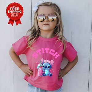 K&ouml;nnte beinhalten: Ein pinkfarbenes T-Shirt mit dem Wort "Stitch" in rosa Buchstaben und einem Cartoon-Bild von Stitch mit einem Getr&auml;nk. Das T-Shirt wird von einem Kind getragen, das eine Sonnenbrille und eine wei&szlig;e Schleife tr&auml;gt.