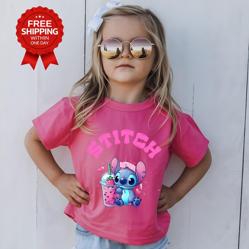 K&ouml;nnte beinhalten: Ein pinkfarbenes T-Shirt mit dem Wort "Stitch" in rosa Buchstaben und einem Cartoon-Bild von Stitch mit einem Getr&auml;nk. Das T-Shirt wird von einem Kind getragen, das eine Sonnenbrille und eine wei&szlig;e Schleife tr&auml;gt.