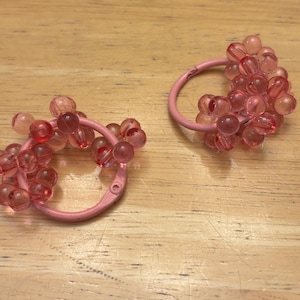 May include: A pair of circular hair accessories featuring a light pink metal ring base adorned with clusters of translucent, round, pink beads. The beads are arranged in a dense, textured pattern around the circumference of each circular frame.