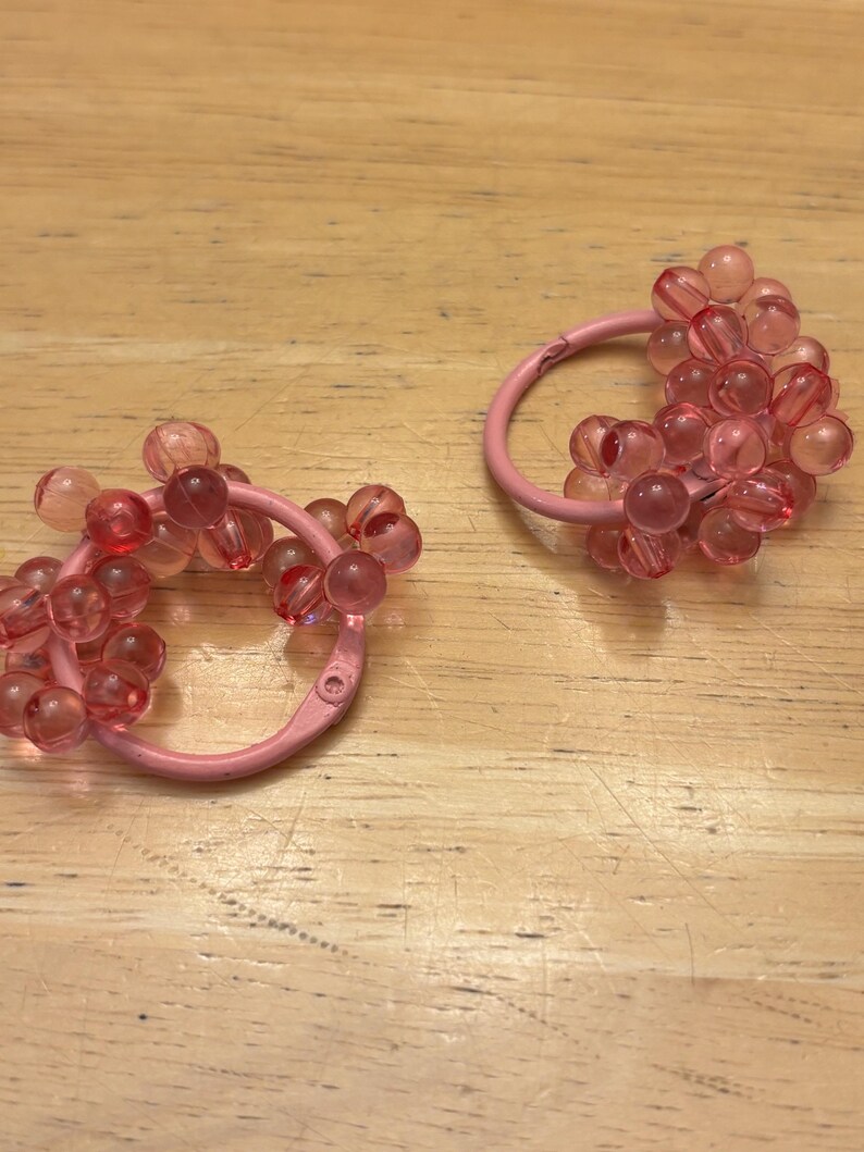 May include: A pair of circular hair accessories featuring a light pink metal ring base adorned with clusters of translucent, round, pink beads. The beads are arranged in a dense, textured pattern around the circumference of each circular frame.