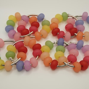 May include: A collection of colorful beaded bracelets. Each bracelet features a series of matte finish, round beads in a rainbow of colors, strung on a silver-tone metal ring. The beads include shades of red, orange, yellow, green, pink, and purple.