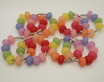 May include: A collection of colorful beaded bracelets. Each bracelet features a series of matte finish, round beads in a rainbow of colors, strung on a silver-tone metal ring. The beads include shades of red, orange, yellow, green, pink, and purple.