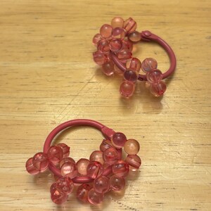 May include: A pair of hoop earrings featuring a matte pink metal circular frame adorned with clusters of translucent, light pink spherical beads. The beads are arranged in small groups along the lower portion of each hoop, creating a textured, layered appearance with a soft, glossy finish.