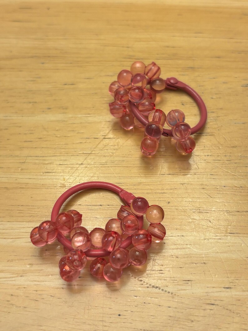 May include: A pair of hoop earrings featuring a matte pink metal circular frame adorned with clusters of translucent, light pink spherical beads. The beads are arranged in small groups along the lower portion of each hoop, creating a textured, layered appearance with a soft, glossy finish.