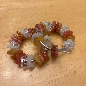 May include: A pair of hoop earrings featuring a series of small, flower-shaped beads in shades of translucent white, clear, amber, and reddish-orange. The beads are strung onto a thin, circular silver-toned metal wire frame that forms the hoop.