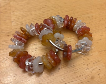 May include: A pair of hoop earrings featuring a series of small, flower-shaped beads in shades of translucent white, clear, amber, and reddish-orange. The beads are strung onto a thin, circular silver-toned metal wire frame that forms the hoop.