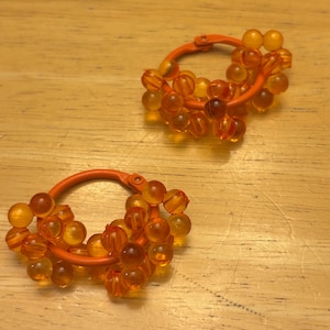 May include: A pair of hoop earrings featuring a solid orange circular frame adorned with multiple translucent orange spherical beads clustered around the exterior. The beads have a smooth, glossy finish and are arranged in a dense, layered pattern.