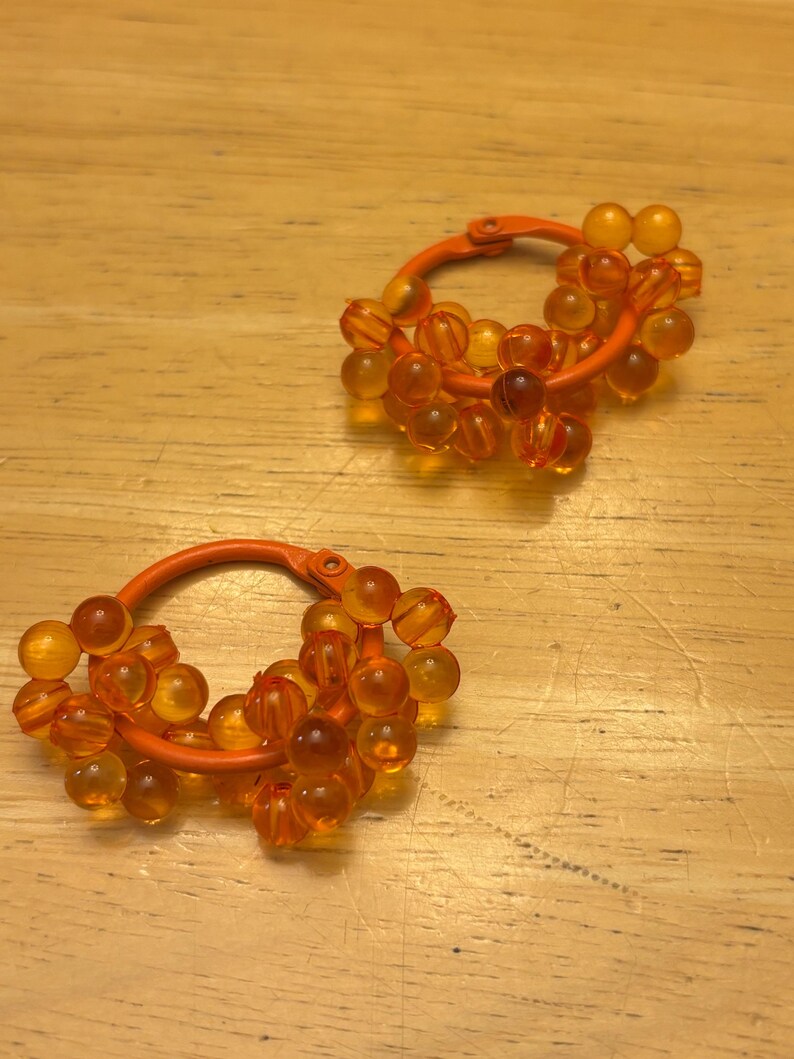 May include: A pair of hoop earrings featuring a solid orange circular frame adorned with multiple translucent orange spherical beads clustered around the exterior. The beads have a smooth, glossy finish and are arranged in a dense, layered pattern.